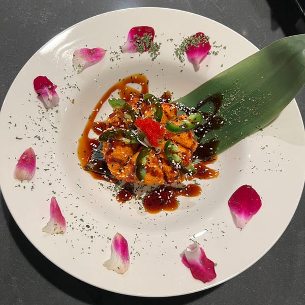 Spicy orange dish with jalapeños, red roe, sesame seeds, and sauce, garnished with rose petals.