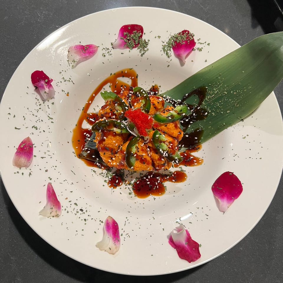 Spicy orange dish with jalapeños, red roe, sesame seeds, and sauce, garnished with rose petals.