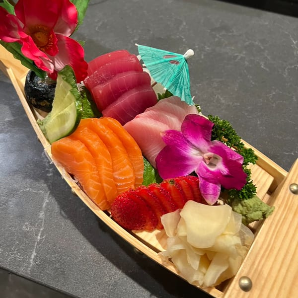 A wooden boat filled with salmon and tuna sashimi, garnished with flowers, strawberries, and ginger.
