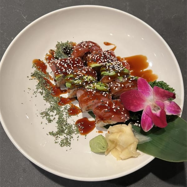 Dragon Roll sushi with avocado, tuna, sesame seeds, sauce, and a pink orchid on a white plate.