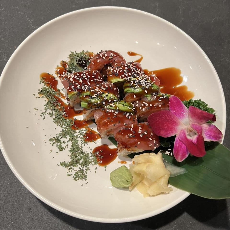 Dragon Roll sushi with avocado, tuna, sesame seeds, sauce, and a pink orchid on a white plate.